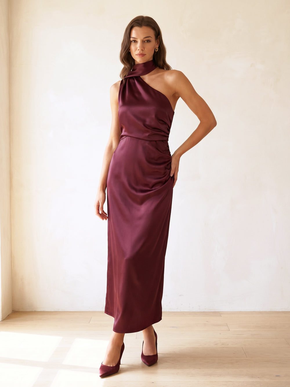 Woman in a wine one shoulder satin maxi dress standing in a sunlit ivory room